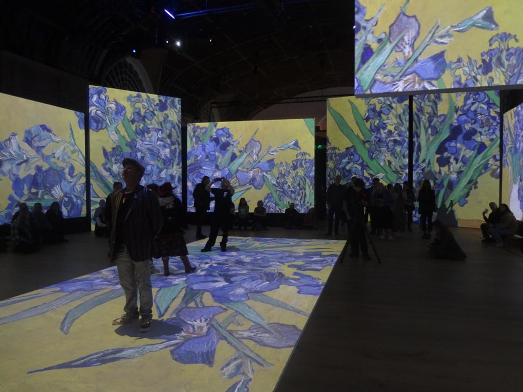 Group of people standing around room, watching projected images of of Van Gogh's art on walls and floor