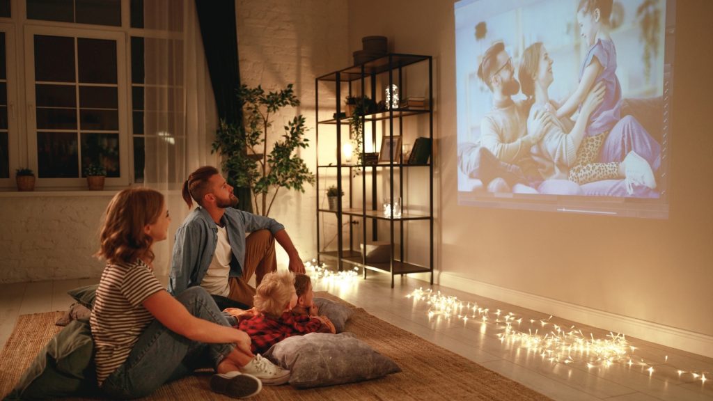 Family made up of a mom, dad and son sitting on the floor on pillows and looking at a movie being projected onto the wall. There are fairy lights on the floor.