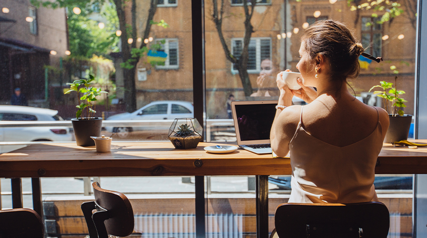 girl sitting at window of cafe with a coffee cup in her hand and laptop on the table looking out onto the road ahead with cars, trees and buildings.