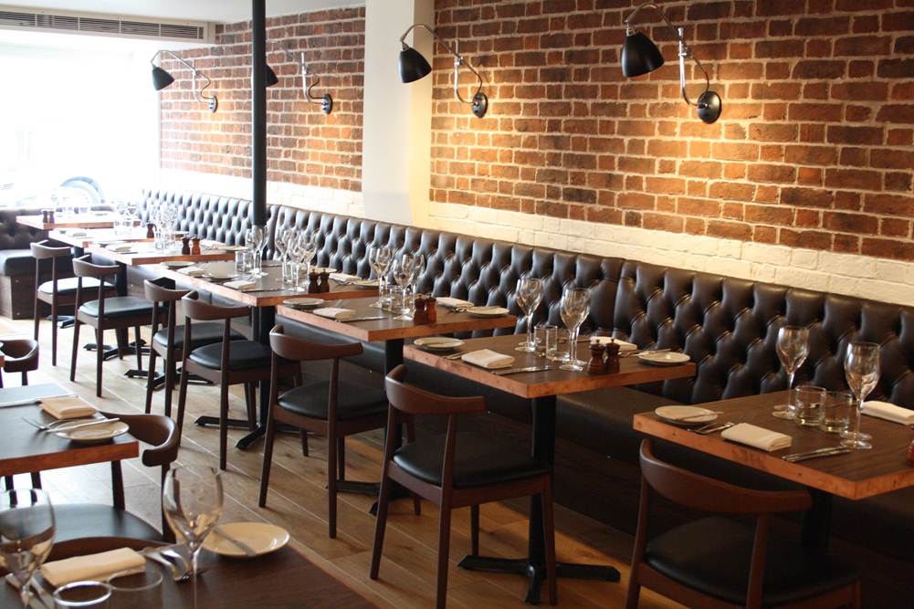 room with brick walls featuring wooden tables with wine glasses, napkins and cutlery, with cushion seats on one side and chairs on other