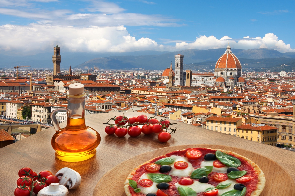 view of italian architecture below a blue sky with a table featuring a pizza, oil and toamtoes.