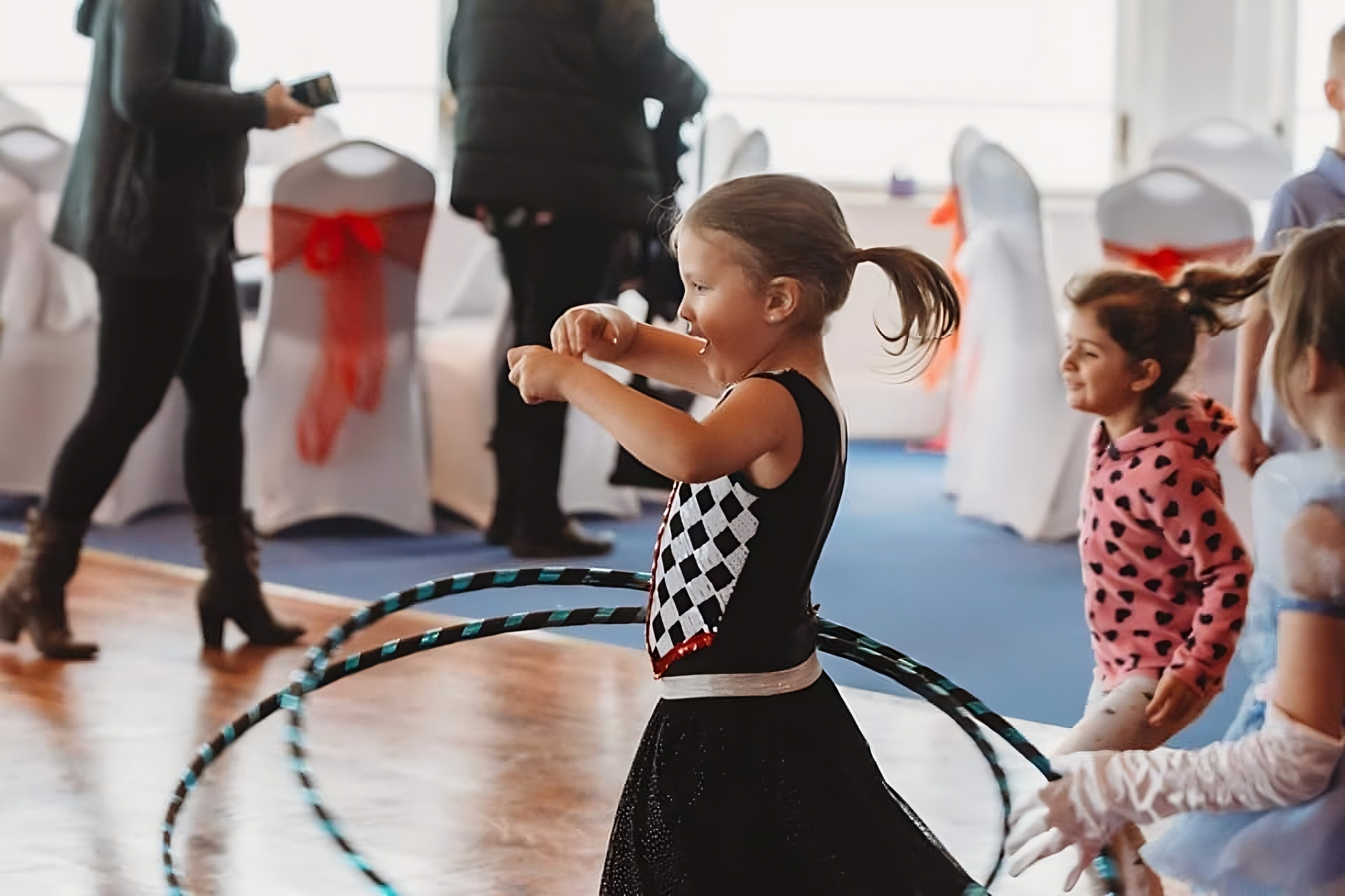 Little girl dressed in a black skirt and black and white top hula-hooping in the middle of a room with some other people around
