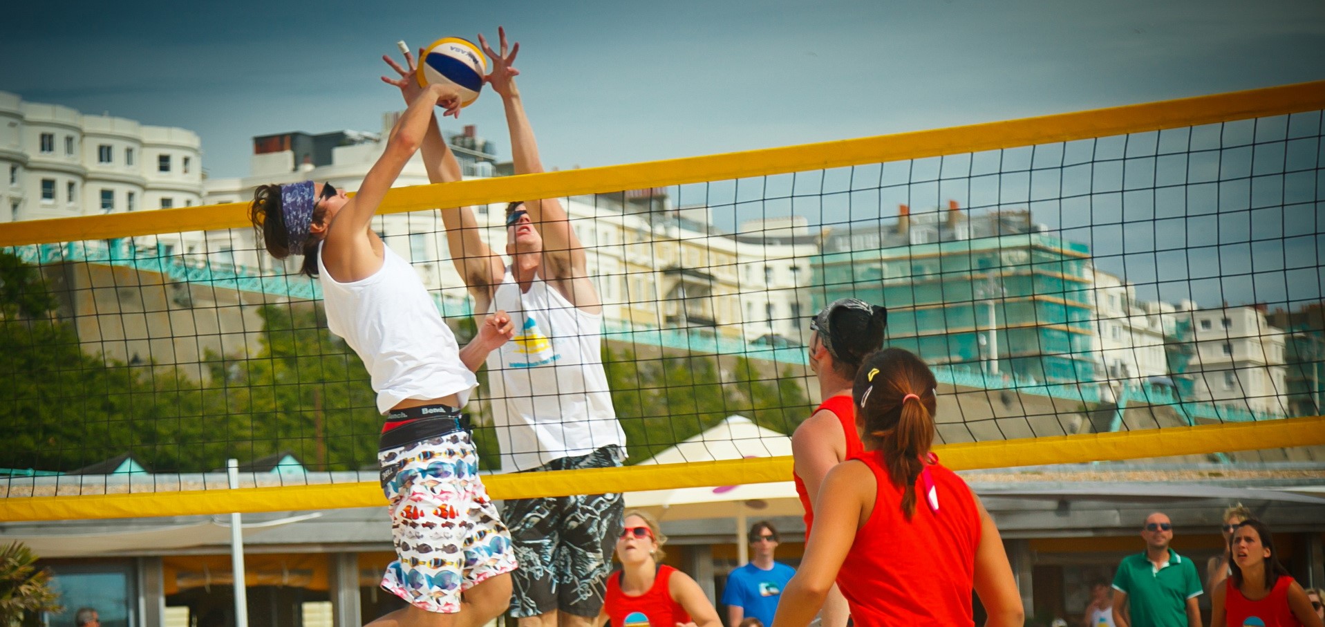 two men jumping on either side of net to reach a volleyball with two girls in red swimsuits watching