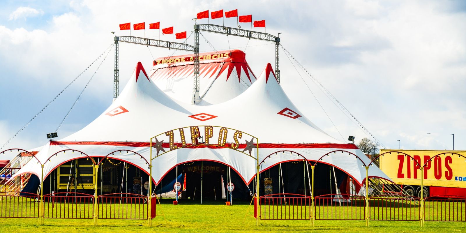 Big white tent with red elements and flags with a fence in front which has ZIPPO in yellow writing