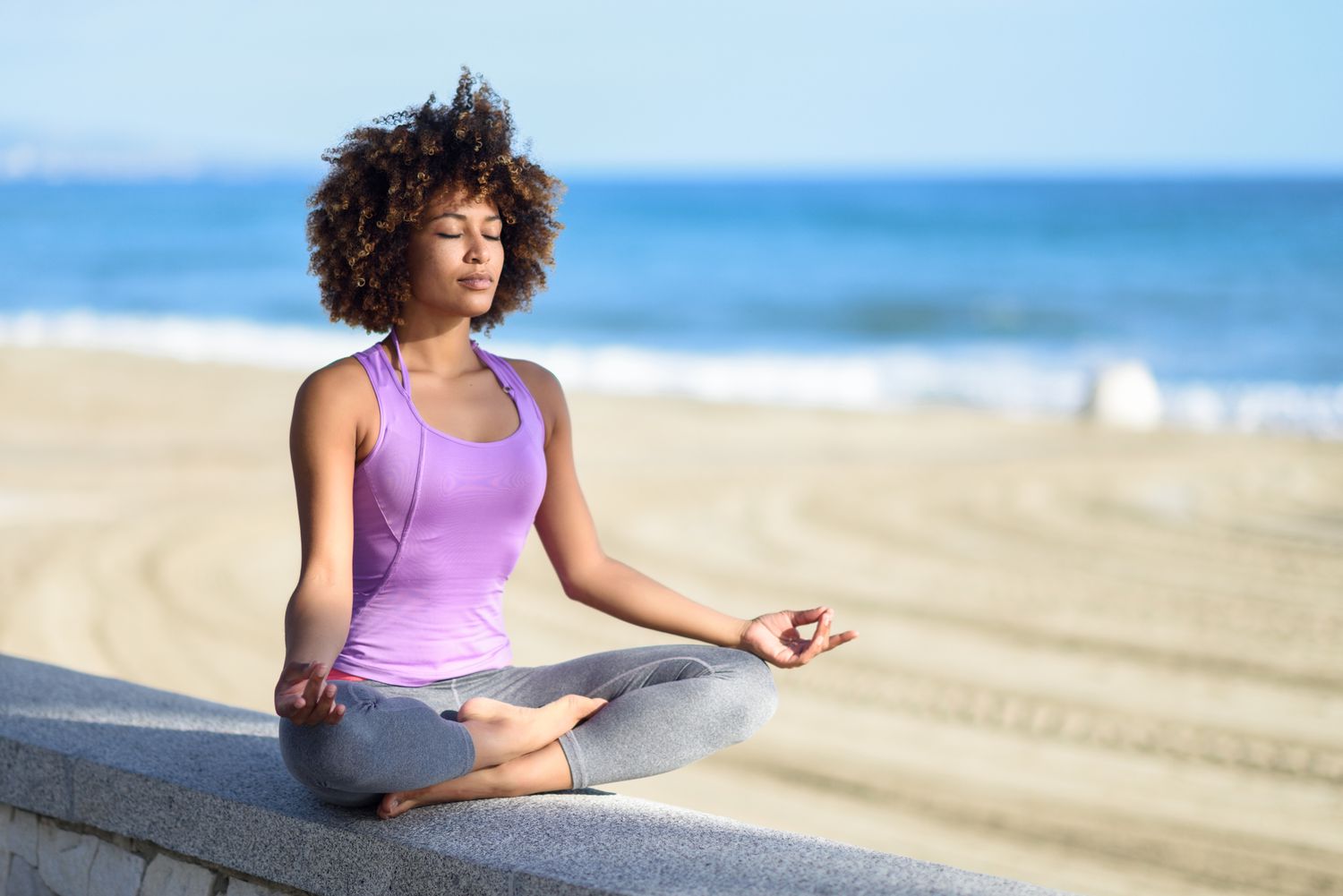 Woman in purple shirt sitting on a beach cross legged with her hands on her thighs and with her eyes closed.