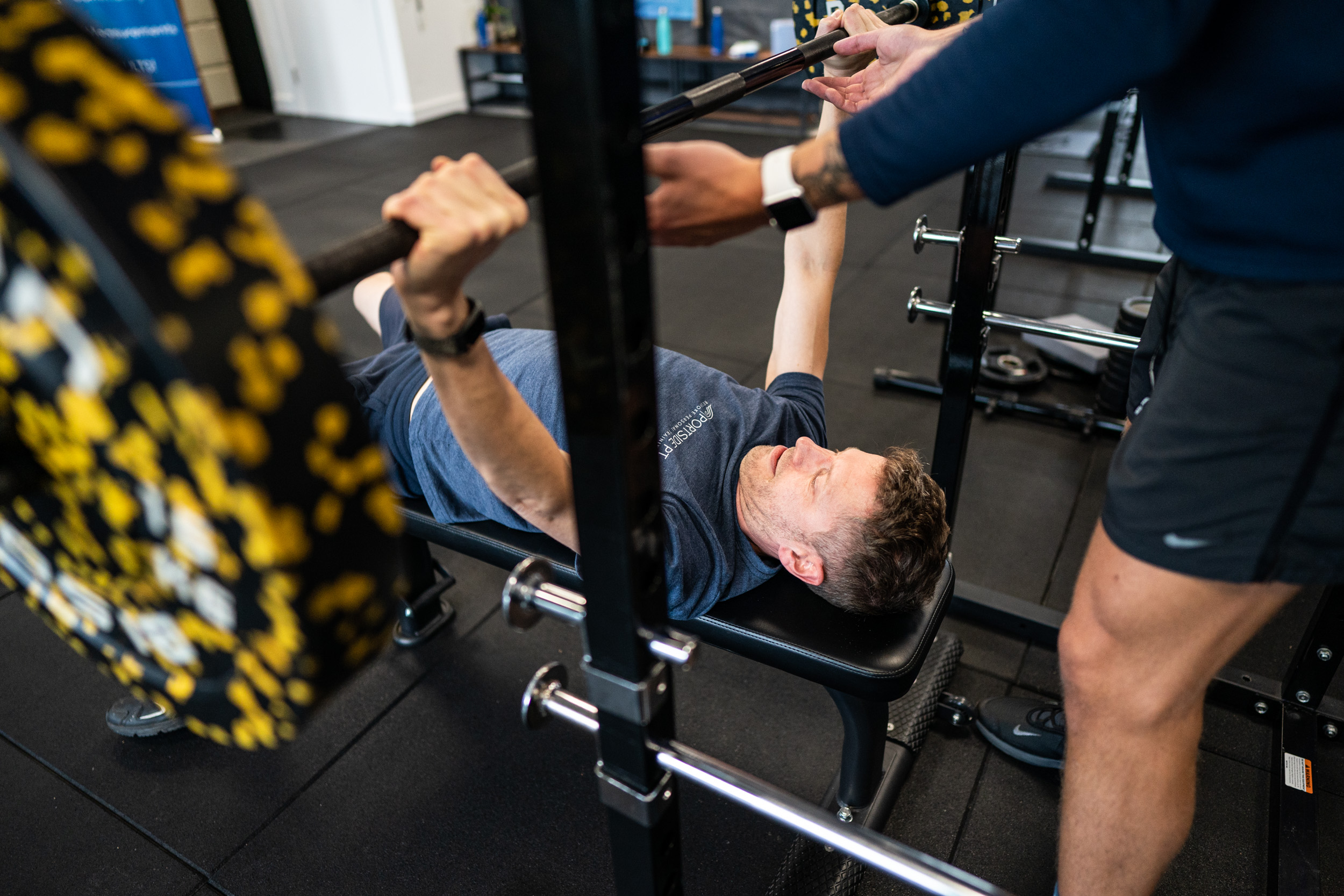 A man laying down on a workout bench lifting a weight with another man standing behind him holding the weight to help him out.