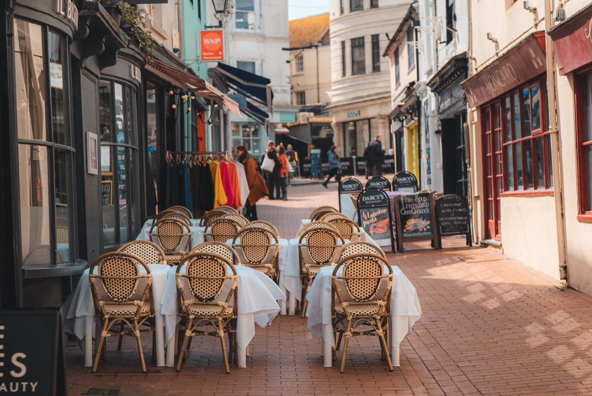 A narrow street with a range of shops and restaurants. The image is featuring wooden chairs and tables with white cloths set up for outdoor seating at a restaurant.