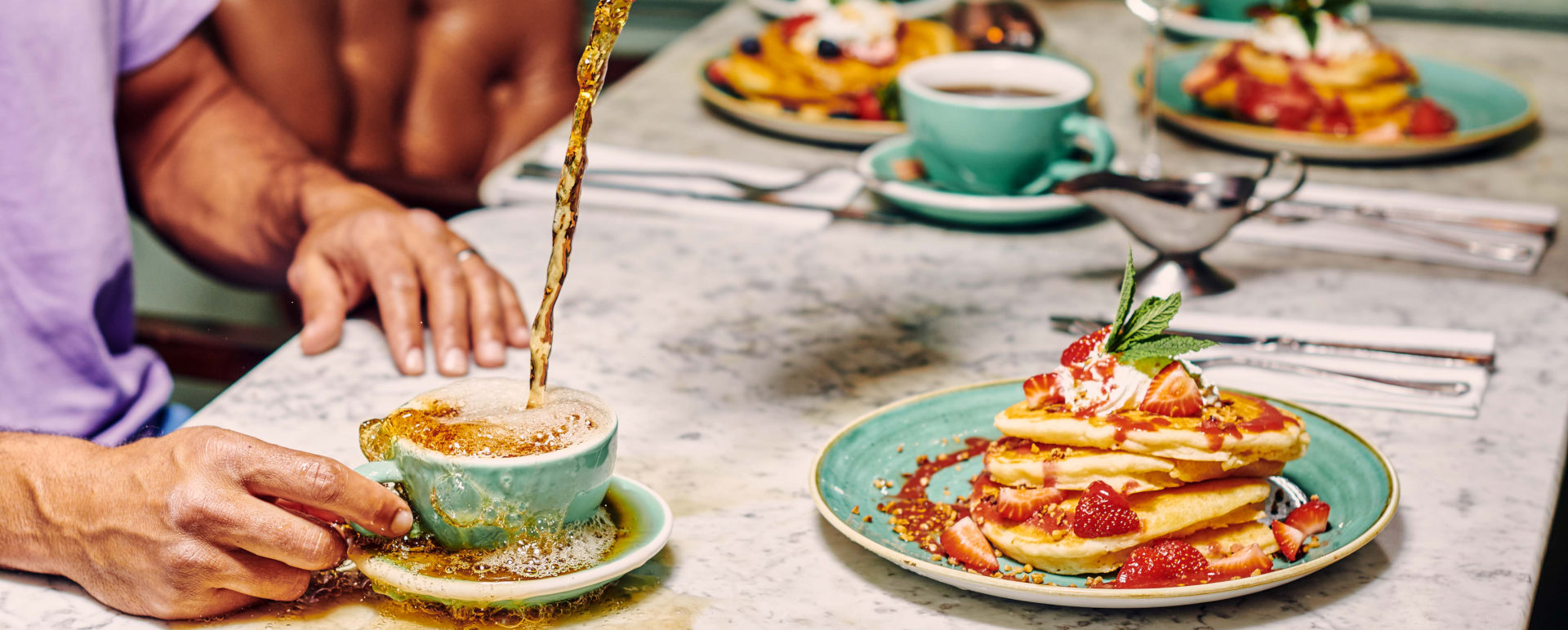 table set with green plates with pancakes drizzled in sauce and with strawberries. Someone is pouring tea from a red teapot into a teacup which is overflowing.
