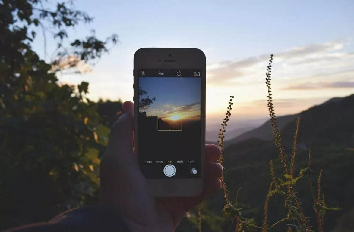 Someone holding up a phone with the camera app open and taking a picture of the scenery which is a sunset over some hills.