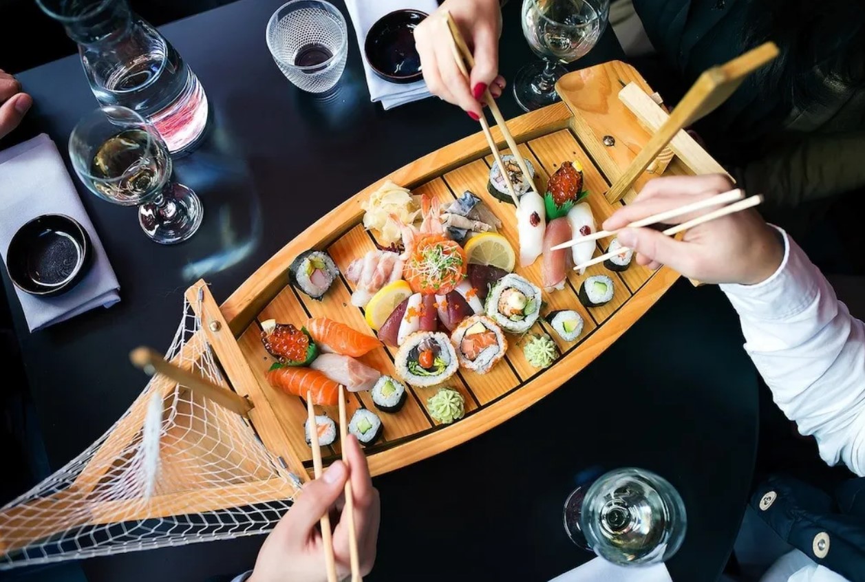 A table with drinks and a wooden boat shaped plate with a range of sushi. People are picking up sushi with chopsticks.