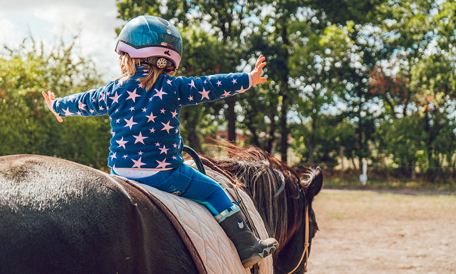 little girl wearing a blue jumper with pink stars sitting on a brown horse with her arms out