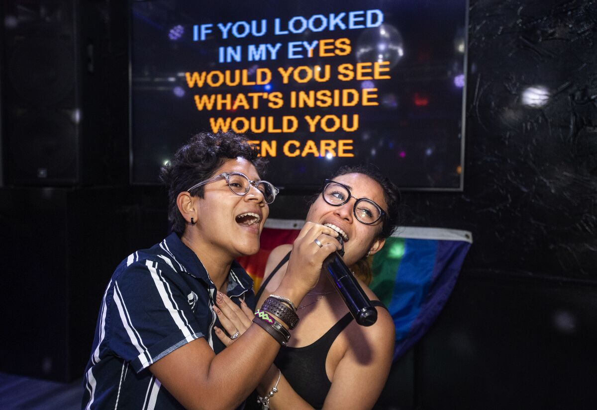Two people singing into a microphone with a screen behind them showing lyrics in blue and orange