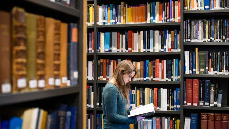 Girl in blue jumper reading a book among the shelves in a library filled with a range of books of different colours