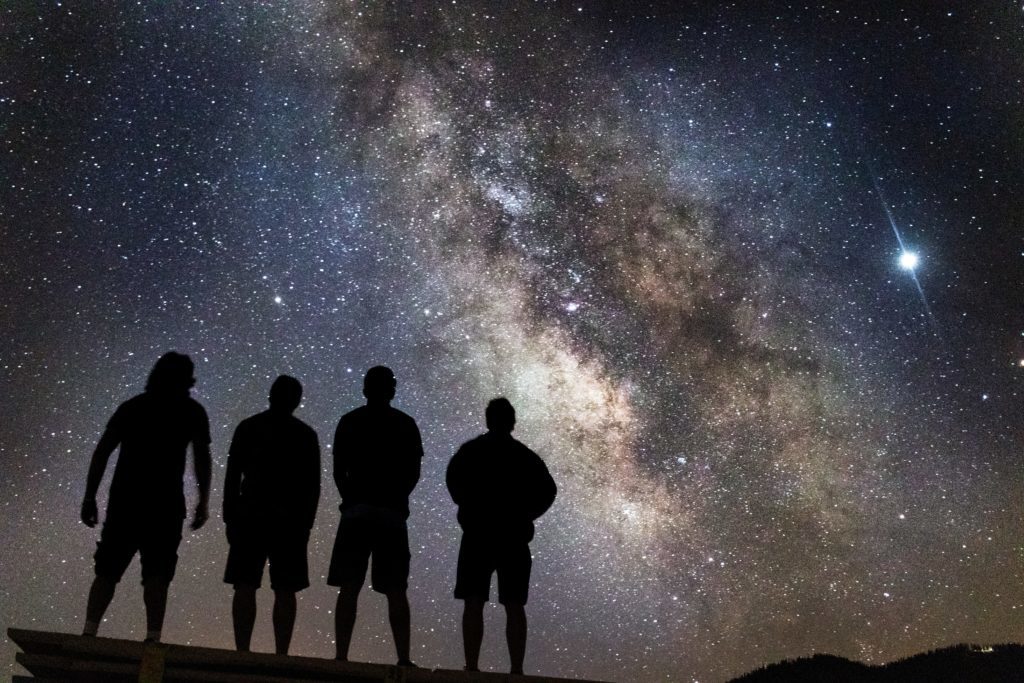 A group of 4 people standing and gazing at the sky which is filled with stars and a milky way.