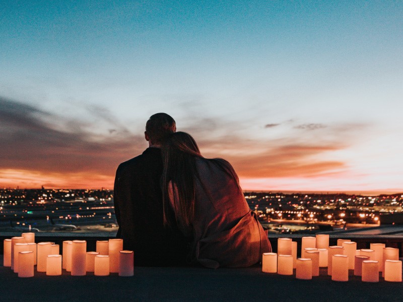 A man and woman sitting with their backs facing to the camera, the woman is leaning her her on the mans shoulder as they admire the view of the sunset and buildings ahead. they are surrounded by lit candles.