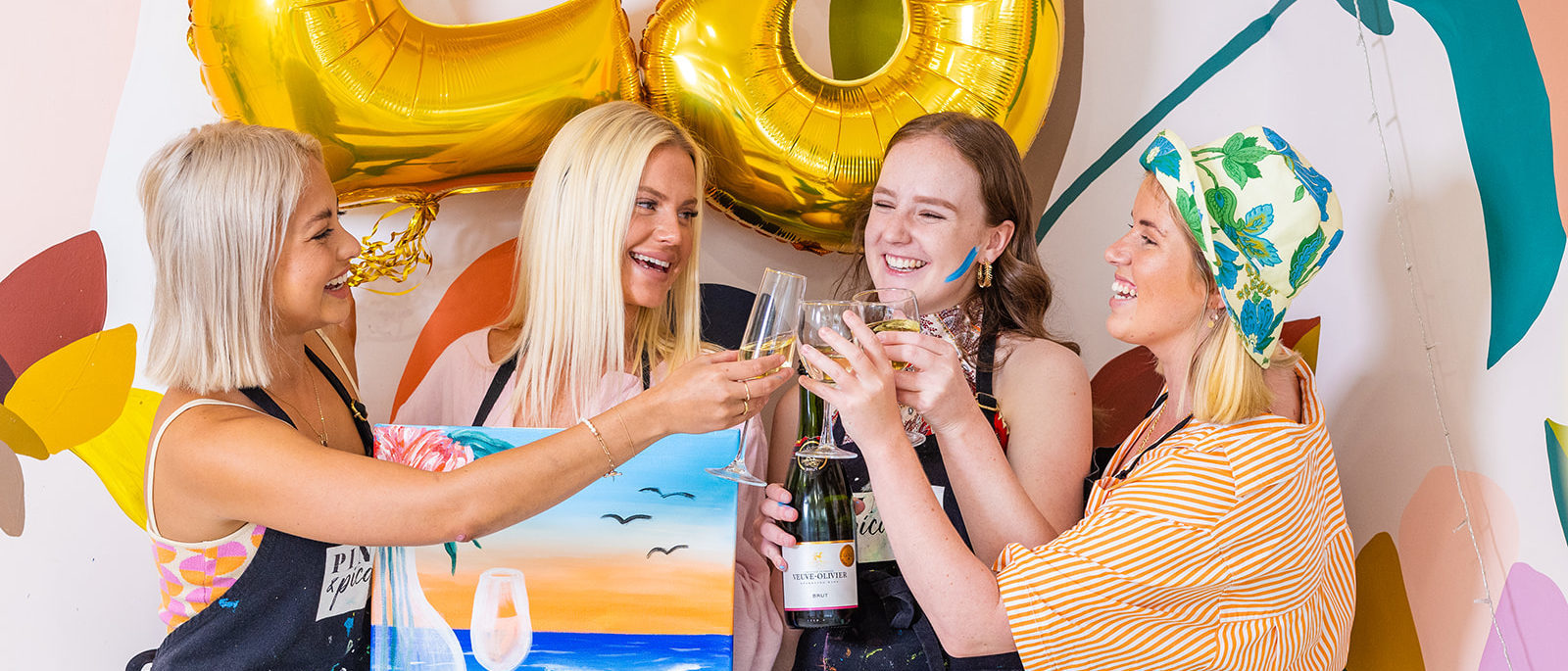 four girls standing in front of a 23 balloon with wine glasses and holding a painting