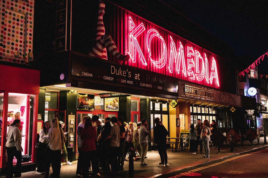 The outside of a building, it is coloured black and features a statue of legs in white and red stripped socks. The building features a sign that says "Duke's at Komedia"