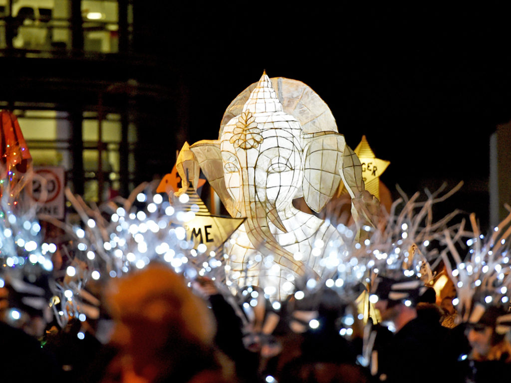 people walking through city centre with lanterns in celebration of burning of the clocks in Brighton. The image features a lanterns shaped like an elephant.