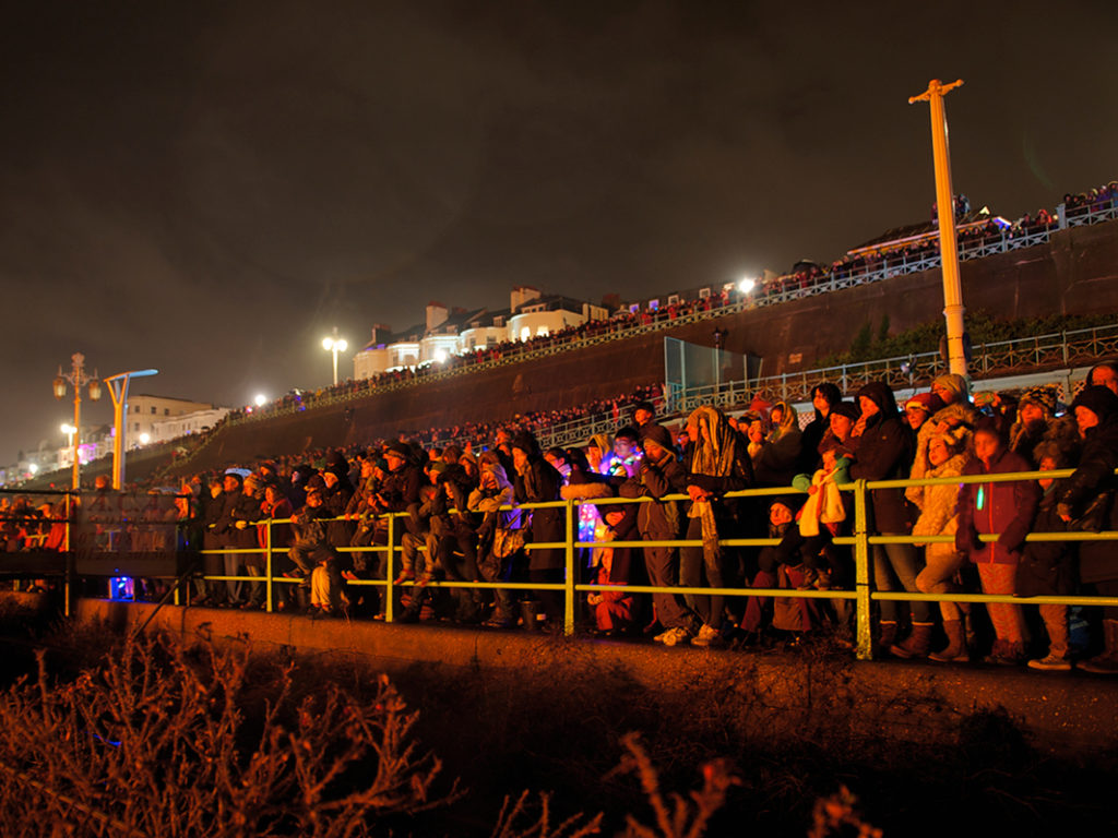 people standing at madeira drive in Brighton watching the fire show on the beach which is part of the burning of the clocks celebration in Brighton